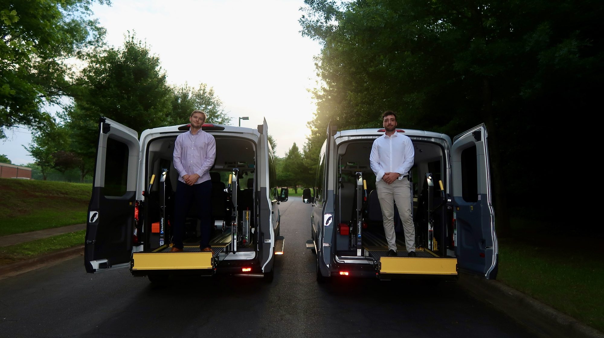 Ray and Mac standing on the wheelchair lifts of two Doctor Ride NEMT vans at sunset
